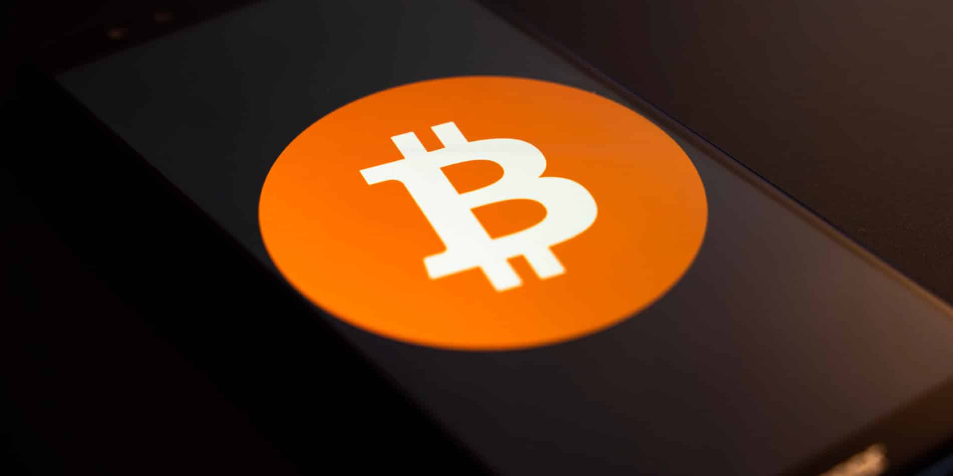 Auf einem Smartphone-Bildschirm wird das Bitcoin-Logo angezeigt. Es besteht aus einem weißen „B“ mit zwei vertikalen Linien, zentriert in einem orangefarbenen Kreis. Das Bild wird schräg auf einem dunklen Hintergrund angezeigt.