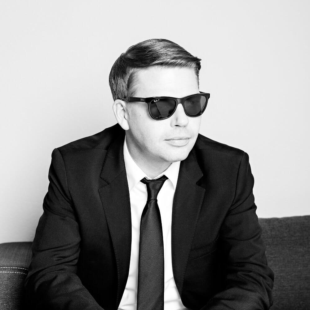 TREGxRM Ein Mann in einem dunklen Anzug, mit Krawatte und Sonnenbrille sitzt auf einer Couch und schaut leicht zur Seite. Das Bild ist schwarz-weiß, und der Mann hat kurzes, ordentlich frisiertes Haar.